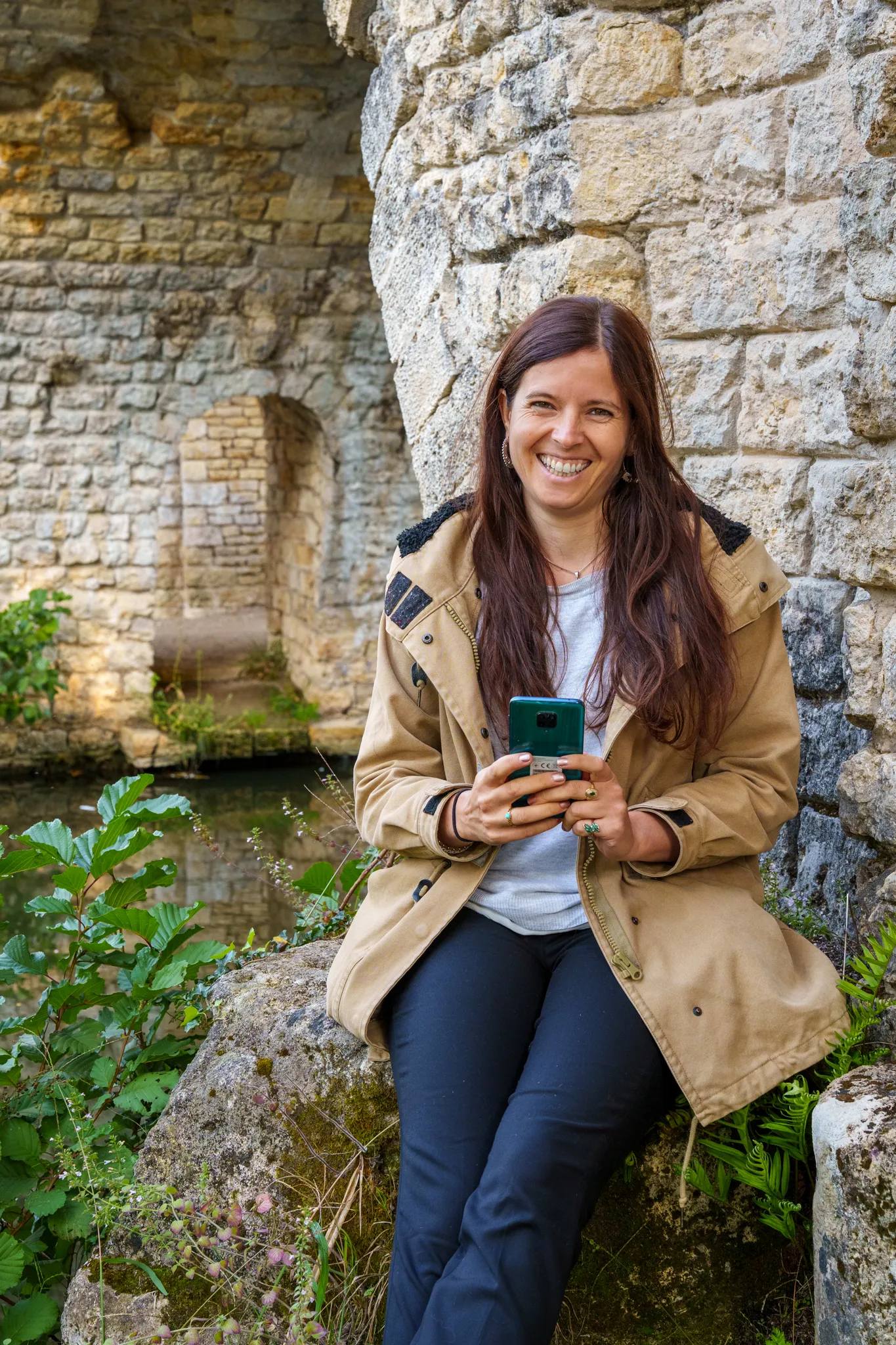 Femme brune cheveux long avec son telephone, près d'un mur en pierre.