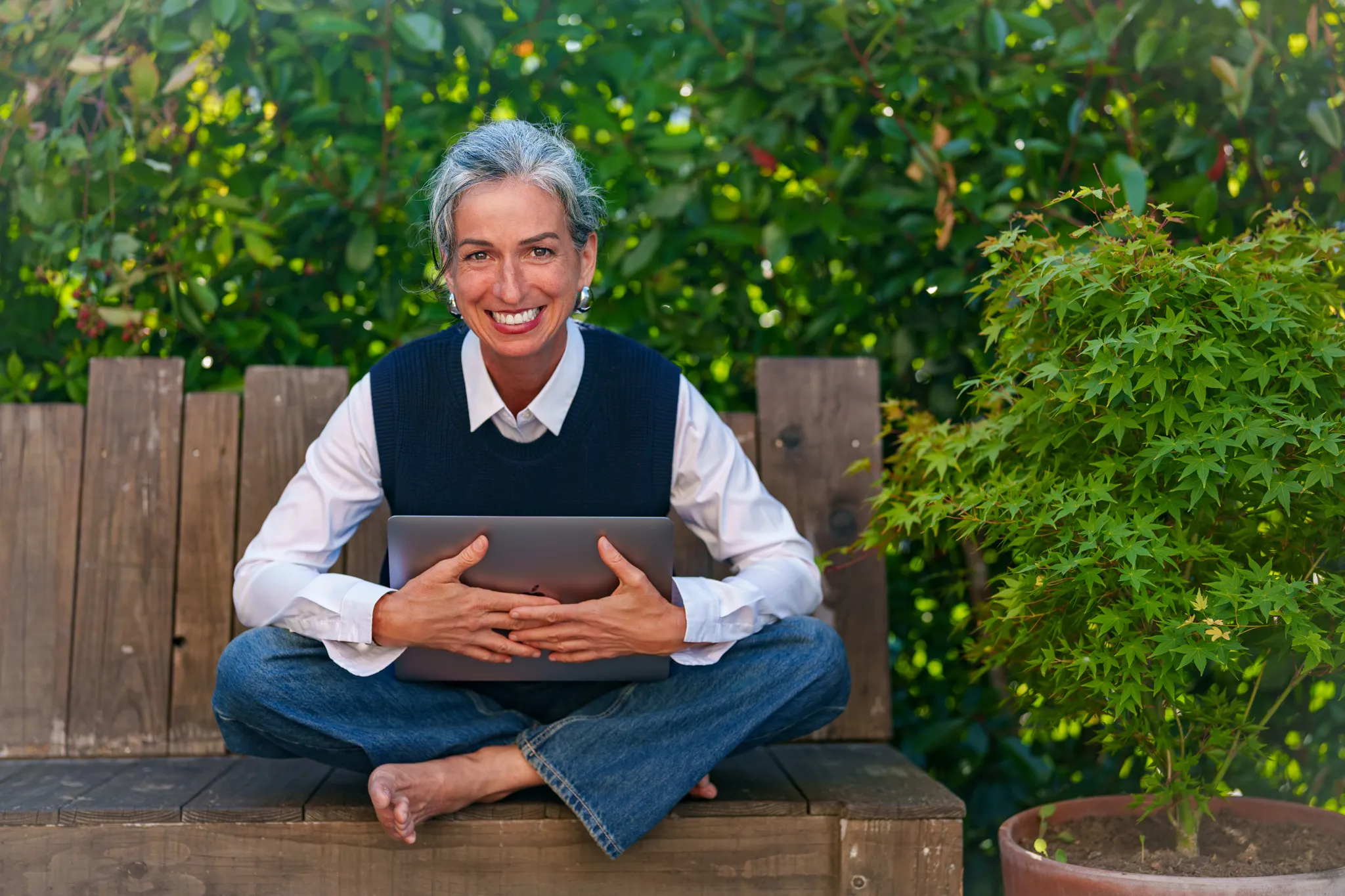 Cheffe d'entreprise, assise en tailleur sur un banc en bois, tenant un ordinateur.